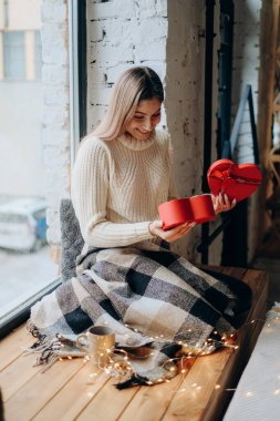 Young beautiful blonde opens a gift for Valentine's Day. A woman opens a red box in the form of a heart with a gift.