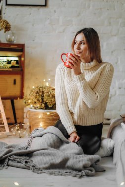 Genç ve güzel bir kadın, Noel arifesinde pencerenin yanındaki bir evde elinde bir kupa sıcak çayla oturuyor..