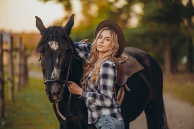 Atı olan genç bir kadın sonbaharda günbatımında çiftliğinde çitin yanında duruyor. Binicilik sporu için safkan atlar yetiştiriyor..
