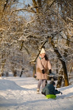 Anne, güneşli kış ormanlarında, karlı patika boyunca plastik kızakla çocuğu çekiyor. Aile aktiviteleri ve boş zaman aktiviteleri..