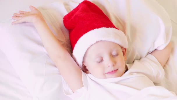 Menino de chapéu de Pai Natal dorme numa cama branca. Ano Novo, feriados, Natal. Manhã felicidade, acorda antes do Natal, véspera de Natal. — Vídeo de Stock