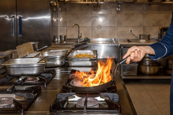 Flame in the pan. Professional Chef makes flambe for food in the restaurant kitchen. Chef cooking with open fire pan on a stove.
