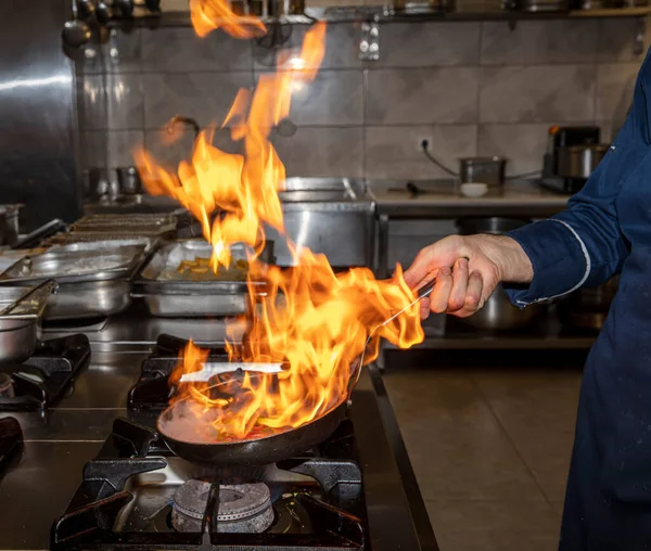 Flame in the pan. Professional Chef makes flambe for food in the restaurant kitchen. Chef cooking with open fire pan on a stove.
