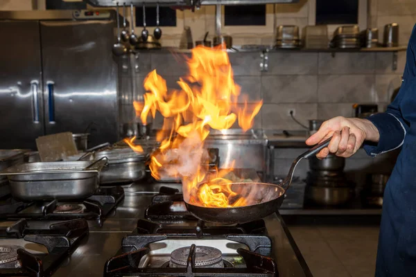 Flame in the pan. Professional Chef makes flambe for food in the restaurant kitchen. Chef cooking with open fire pan on a stove.