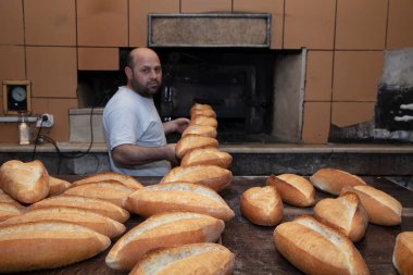 Fırıncı. Genç, yakışıklı bir fırın işçisi ekmek arka planında, sobadan tahta bir kürekle ekmek alıyor. Fırın ürünlerinin endüstriyel üretimi. Fırıncının özel giysileri içinde bir adam. Fırın