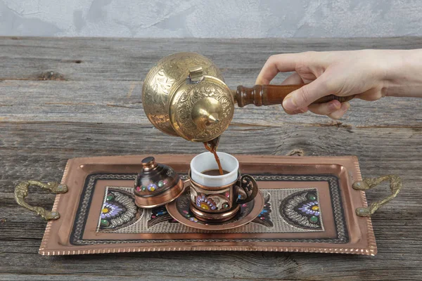 Turkish coffee. Pouring Turkish coffee into vintage cup on wooden background. Pouring Turkish coffee into traditional embossed metal cup.