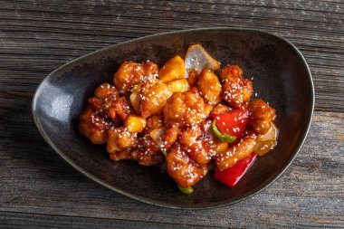 Mançurya usulü kızarmış tavuk parçaları, dolmalık biber ve soğanla pişirilmiş ve koyu baharatlı sosla kaplanmış. Asya 'dan esinlenilmiş Hint-Çin mutfağı karanlık arka planda, yakın plan yemek fotoğrafçılığında modern bir kâsede servis edilir..