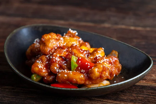Mançurya usulü kızarmış tavuk parçaları, dolmalık biber ve soğanla pişirilmiş ve koyu baharatlı sosla kaplanmış. Asya 'dan esinlenilmiş Hint-Çin mutfağı karanlık arka planda, yakın plan yemek fotoğrafçılığında modern bir kâsede servis edilir..