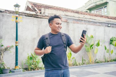 Sırt çantalı gülümseyen Asyalı adam Yogyakarta 'da gezerken cep telefonu kullanıyor.