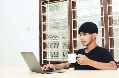 Asyalı bir adam dizüstü bilgisayarın önünde oturup kahve içiyor.