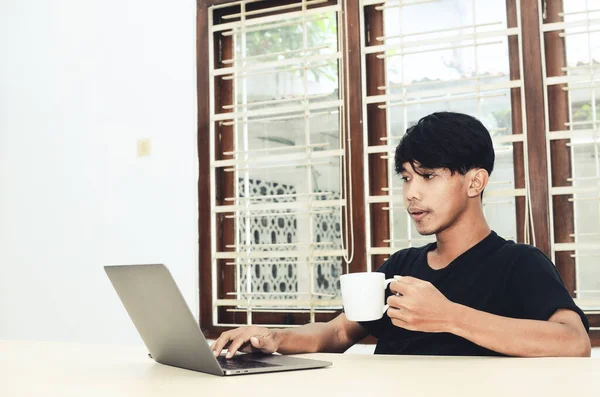 Asyalı bir adam dizüstü bilgisayarın önünde oturup kahve içiyor.
