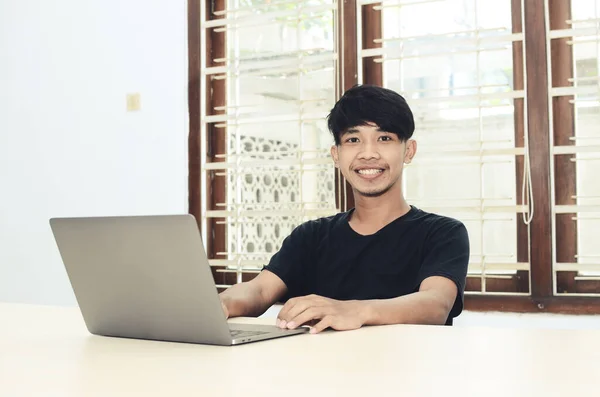 Ofis masasında oturan siyah gömlekli Asyalı adam dizüstü bilgisayarın önünde gülümsüyor.