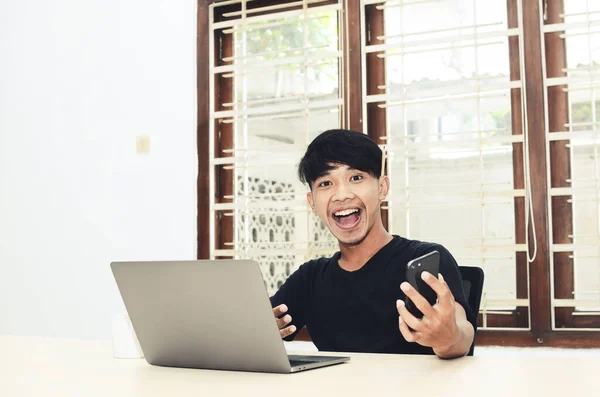 Siyah Asya tişörtlü adam çok mutlu bir ifadeyle dizüstü bilgisayarın önünde oturdu.