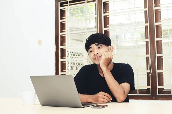 Siyah Asya tişörtlü adam üzgün bir ifadeyle dizüstü bilgisayarın önünde oturdu.