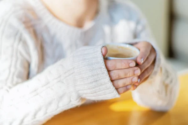 Close-up of female hands in a warm sweater holding a white Cup of hot coffee or tea in a coffee shop. The concept of winter comfort, holiday. The girl warms her hands with a Cup of hot coffee