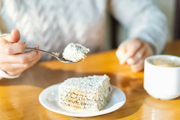 Women\'s hands in a warm knitted sweater in a city cafe with a teaspoon cut off a piece of delicious cake. Coffee mug on the table. Rest in a coffee shop. Close-up, selective focus.