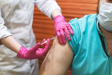 Pembe lastik eldivenli genç bir kadın doktor omzundaki adamı aşılıyor. Yüzünde tıbbi maske olan bir hemşirenin, evde oturup, Corona salgını sırasında bir adama iğne yaptığı yakın plan..