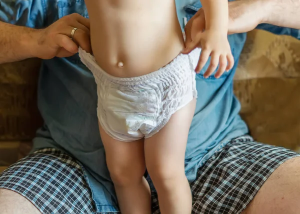A father changes his little daughter\'s diaper while sitting on the couch at home. The hands of a dad changing diapers. A small child in a clean diaper. The concept of parental care. Selective Focus