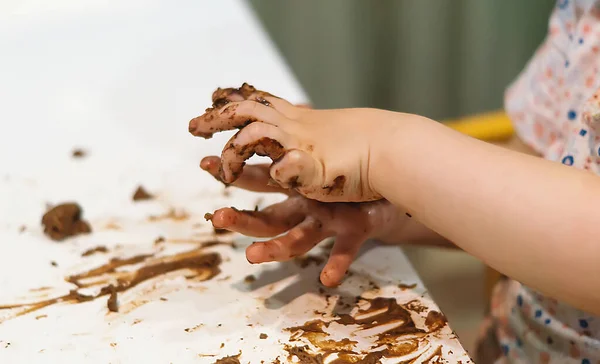 女の子の手は茶色のチョコレートで染色されています 汚い子供の手だ 子供は混乱し キッチンの自宅で白いテーブルの上にチョコレートケーキを掃除しました 幸せな子供時代 ロイヤリティフリーのストック写真