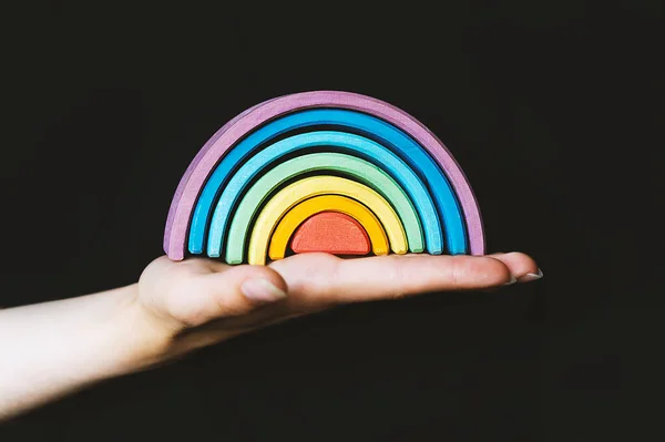 A Hand Holding A Rainbow. A beautiful, multi-colored, wooden rainbow in a woman\'s hand on a black background. A symbol of happiness, love, and hope. Everything will be fine. Banner design.