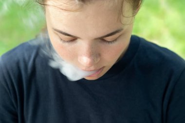 VAPE sigarası. Kahverengi saçlı bir kız yakın planda ağzından buhar salıyor. Sigara içen bir kızın portresi. Sigarayı bırakmak için sigara içiyorum. Sigara konsepti. Kötü bir alışkanlık. Bağımlılık