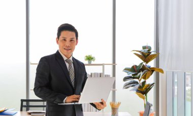 Asyalı bir iş adamı ofiste çalışmak için dizüstü bilgisayarını tutuyor. İş stratejileri için sosyal medya ve online teknoloji haberleri için uygulamaları kullanın. Gülümse ve mutlu ol.