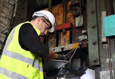 Üniforma ve gözlük takan profesyonel bir erkek mühendis bakım bilgilerini bulmak için dizüstü bilgisayar kullanır. Ve makinelerin endüstriyel fabrikalara yerleştirilmesi mühendislik kavramına güvenerek