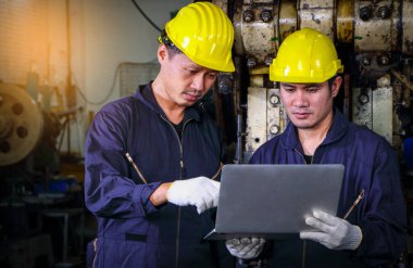 Miğfer üniforması giymiş iki yetenekli Asyalı işçi metal basın denetleme verilerini analiz etmek için bilgisayar kullanıyorlar. Fabrikadaki takım çalışması kavramı.