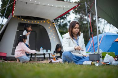 Asyalı anne aile için çadırın dışında yemek pişirirken kamp alanında ailesiyle birlikte mutlu bir şekilde kamp yapıyor. Aile doğa ve kamp kavramının tadını çıkarır..