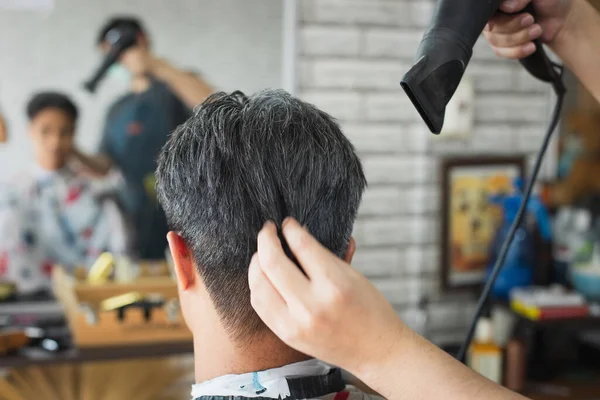 ヘアドレッサーブローヘアカット終了後に彼のクライアントの髪を乾燥させます アジアの若いです男あります打撃乾燥した髪とともにヘアドライヤーによってプロフェッショナル理髪店で — ストック写真