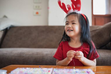 Sevimli Asyalı çocuk Noel kostümü giyip boncukları ipliğe bağlayıp evde eğleniyor..