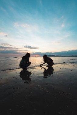 Gün batımında güzel deniz ve gökyüzüyle sahilde oynayan iki Asyalı kız çocuğunun silueti. Aile doğanın tadını çıkarır..