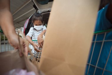 Korunma maskesi takan Asyalı anne ve iki kız Coronavirus salgını sırasında alışveriş arabasının bagajına bir şeyler koymaya yardım ediyorlar..