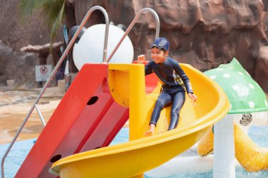 Asyalı bir çocuk yazın su parkında su kaydırağında kayarken eğleniyor..