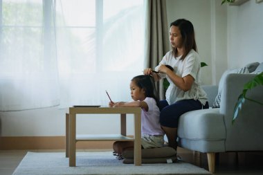 Asyalı çocuk ev ödevi yapıyor ve anne de oturma odasında saçını tarıyor.. 