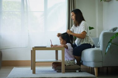 Asyalı çocuk ev ödevi yapıyor ve anne de oturma odasında saçını tarıyor.. 