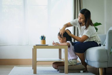 Asyalı çocuk ev ödevi yapıyor ve anne de oturma odasında saçını tarıyor.. 