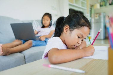 Asyalı kız çocuğu, kız kardeşiyle kanepede oturup öğretmenler ve sınıf arkadaşlarıyla görüntülü konuşma yapıyor. Çocuklar COVID-19 salgını nedeniyle karantina sırasında evde eğitim görüyorlar..