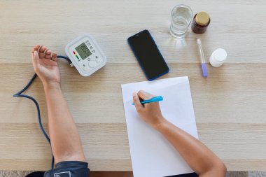 Asyalı genç bir kadın kan basıncını ve kalp atışlarını dijital basınç göstergesi ile kontrol ediyor ve evdeki kağıt üzerinde kayıt yapıyor. Sağlık ve Tıp Konsepti.