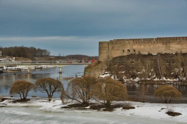 Şehir manzarasında bahar başlarında Narva sınır nehrinin iki kıyısı, Estonya tarafında grimsi ıslak kar üzerinde gür söğüt çalıları ve Rus tarafında antik bir kale yer alıyor..