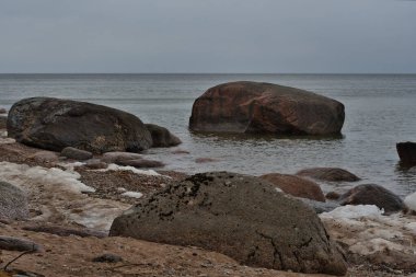 Deniz kenarlarında, kumlu bir kıyıya yakın granit kayalar, kar kalıntıları ve bulutlu bir gökyüzünün altında yağmurlu bir nisan gününde Narva Körfezi 'nin sularında..