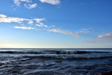 Gün batımından önce akşam denizinde dalgalar mavi bir gökyüzünün altında hafif bulutlarla birlikte, kendileri gökyüzüne gölgeler saçmaya başlarlar..