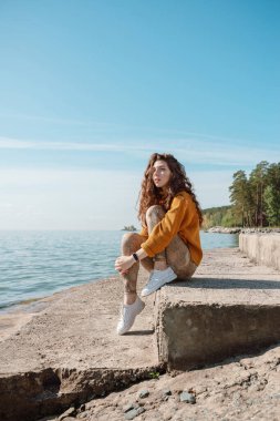 Turuncu kazaklı bir kadın kumsalda taş bir merdivende oturmuş gökyüzüne bakıyor, seçici bir şekilde.