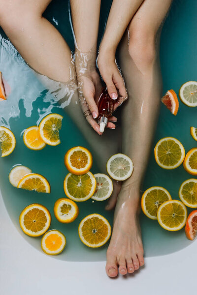 Faceless shot of woman in a bathroom filled with blue water and various cut citrus fruits holding serum in her hands
