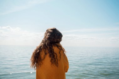 Sahilde, suyun önünde sarı kazaklı, seçici bir odak noktası olan bir kadının arka planı.