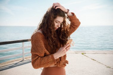 Plajda akıllı telefonunu tutan turuncu kazaklı ve tozlu genç bir kadın, seçici bir şekilde odaklanmış.