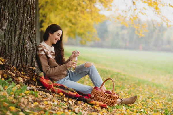 Portrait de belle jeune femme assise près de l'arbre et buvant du cidre d'une bouteille. — Photo