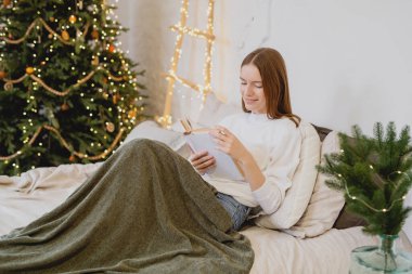 Çekici genç bir kadın, bacaklarında ekoseyle yatakta uzanıp kitap okuyor..