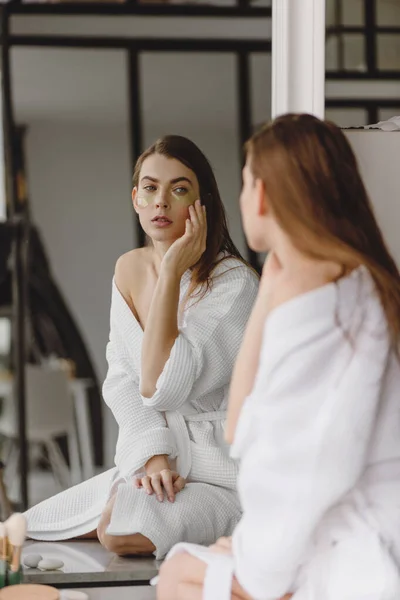 Hermosa mujer en albornoz blanco sentada frente al espejo en el baño y poniendo parches en la cara. —  Fotos de Stock