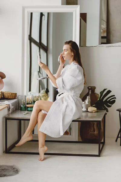 Junge Frau im weißen Bademantel sitzt vor Spiegel und schminkt sich. — Stockfoto